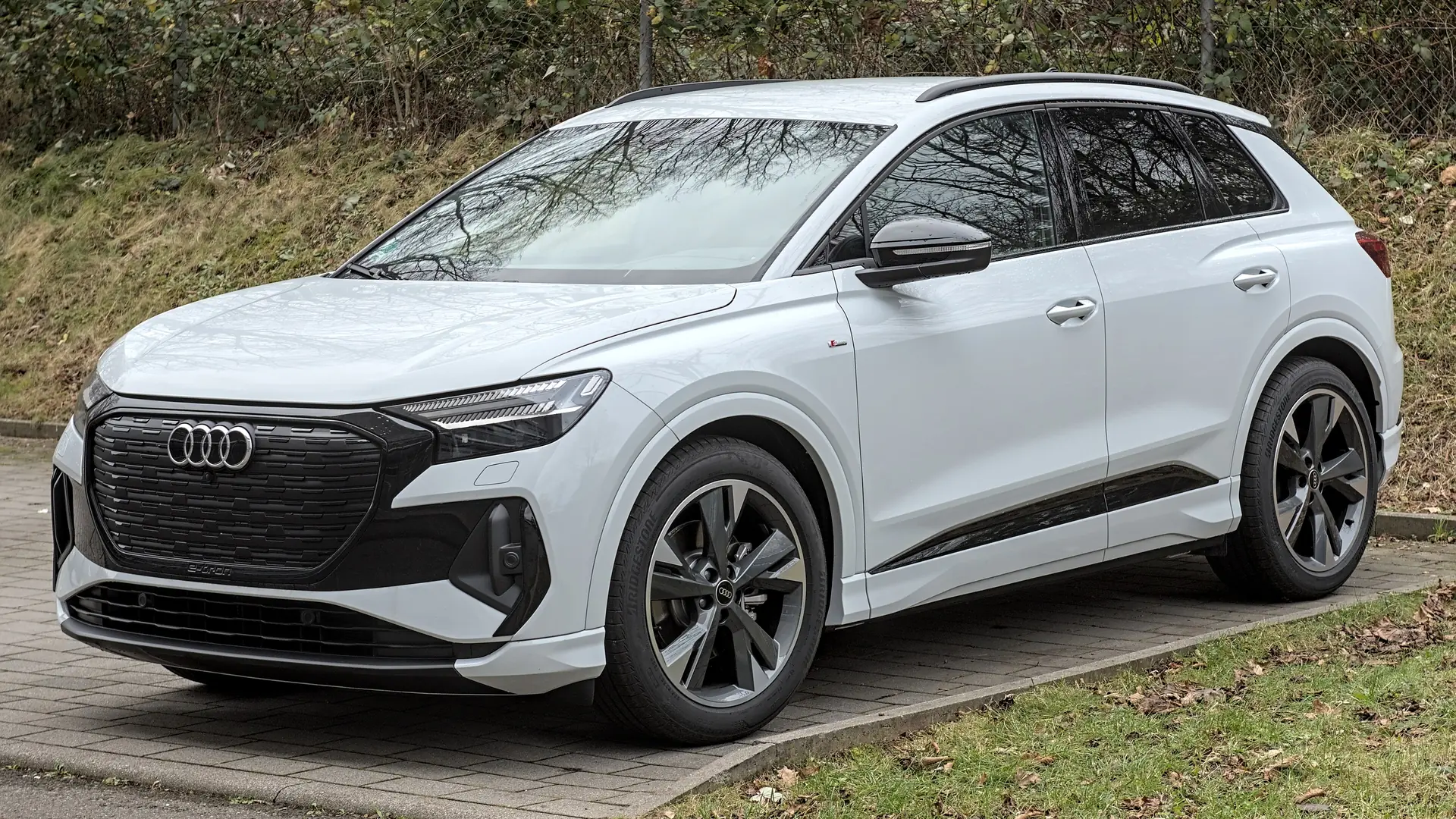 Een witte Audi Q4 e-tron, schuin van voren gezien, geparkeerd op een klinkerstrook voor een grasberm met struiken en een hek