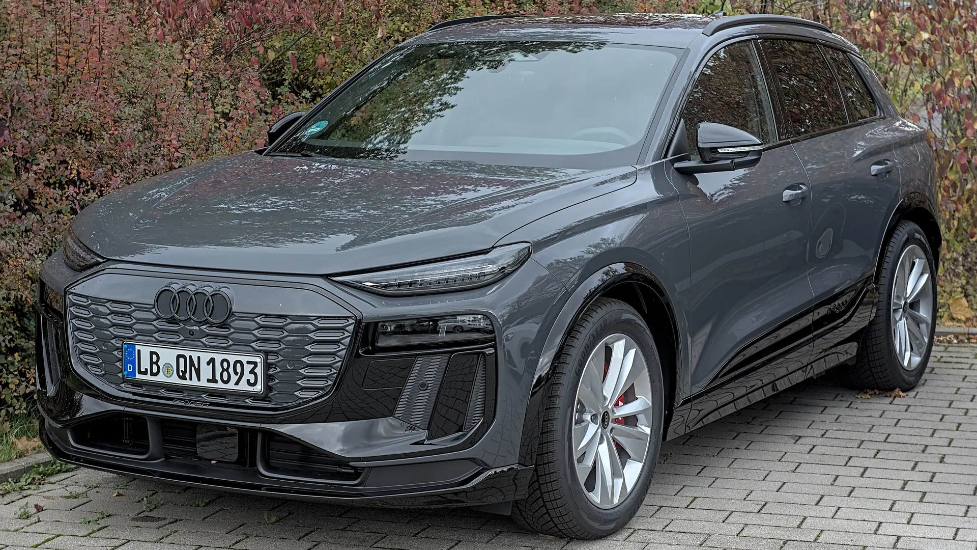 Een grijze Audi Q6 e-tron, schuin van voren gezien, geparkeerd op een klinkerterrein voor een haag met herfstbladeren