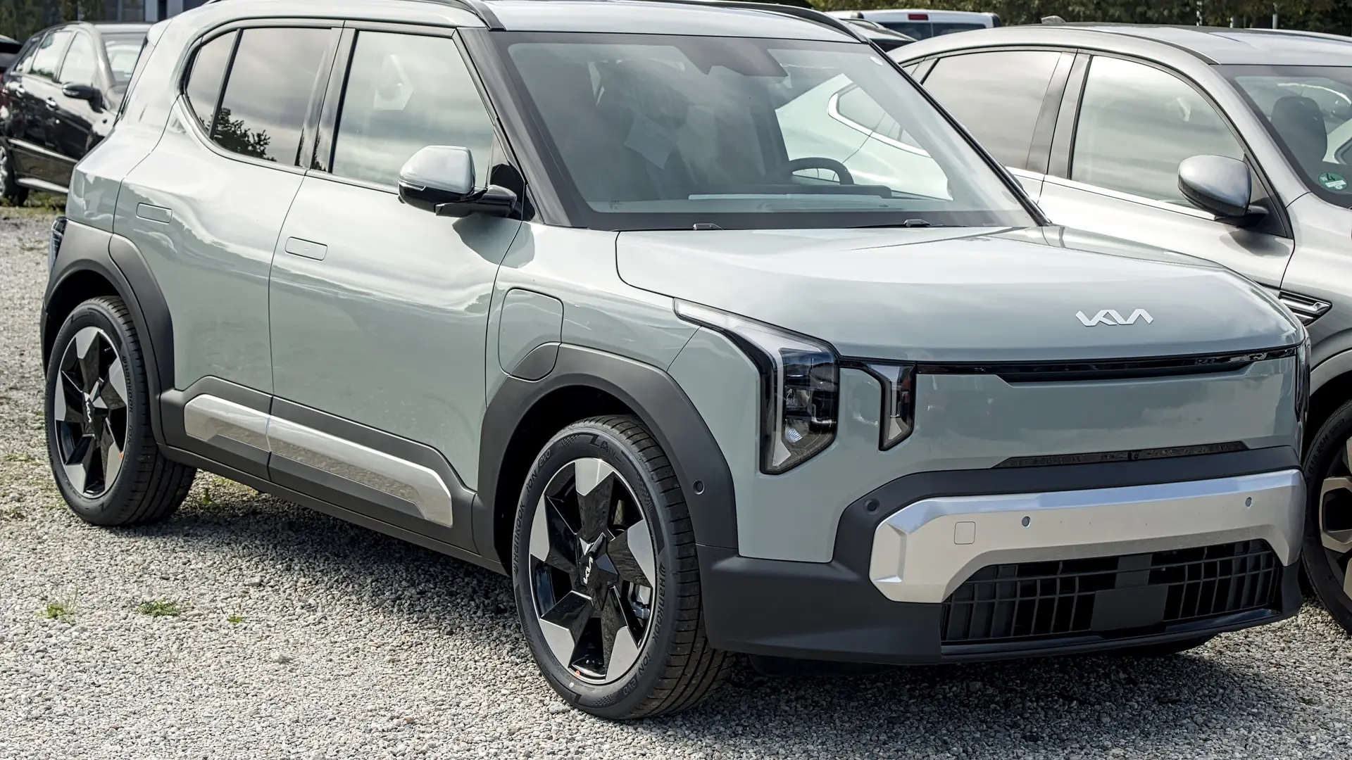 Een lichtgrijze Kia EV2, schuin van voren, geparkeerd tussen andere auto’s