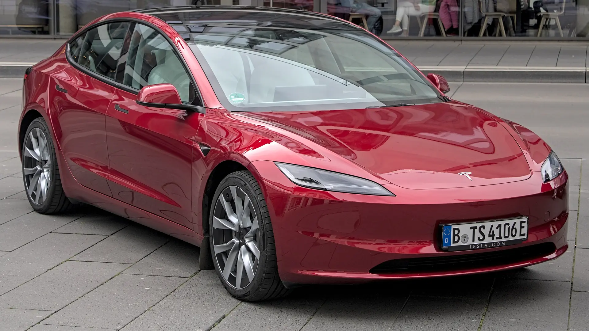 Een rode Tesla Model 3, schuin van voren, op een plein voor een café